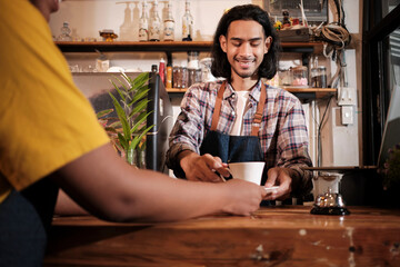 Close-up shot, young long hair Thai male baristas startup works by serving a cup of coffee to a customer with a smile at casual cafe counter bar, happy service job, and small business entrepreneur.