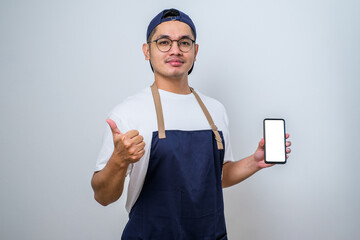 Young Asian handsome barista man wearing apron ands smiling, showing smartphone and thumbs up gesture, looking at camera