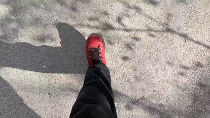 Feet with red shoes walking on grey asphalt pavement on a sunny day POV