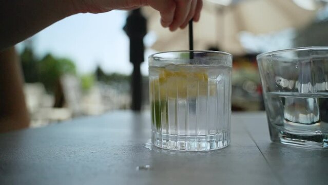 Hand uses a straw to mix a glass of water with ice cubes, lemon and lime in it. Super slow motion.