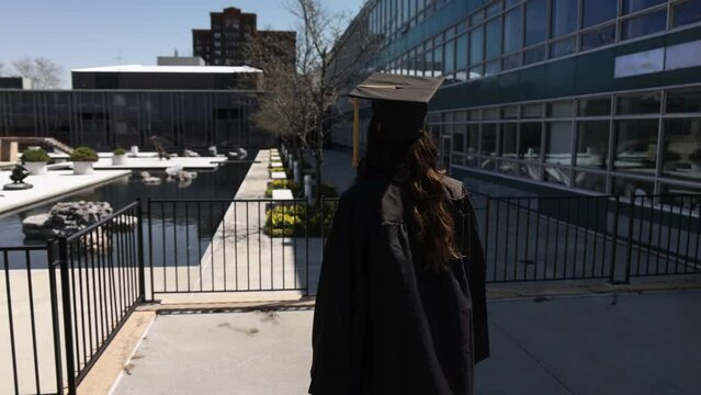 Female college graduate wearing cap and gown walking outside with video from behind in slow motion.
