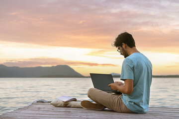 Young digital nomad man sitting on wooden pier at sea working on internet remotely at sunset -...