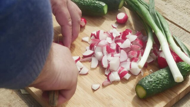 a hand cuts a radish with a knife on a cutting board next to leeks and cucumbers