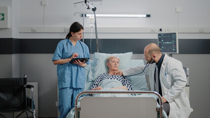Doctor consulting sick patient with stethoscope in hospital ward for heartbeat measurement while...