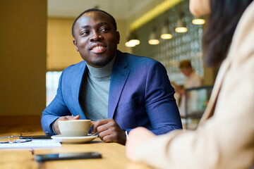 African young businessman in blue suit drinking coffee and talking to his colleague during meeting at cafe