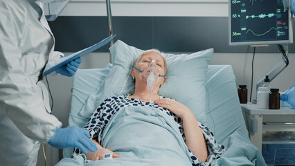 Paramedics consulting heavy breathing woman with oxygen tube in hospital ward quarantine. Doctor and nurse with suits and masks assisting aged patient during coronavirus outbreak.