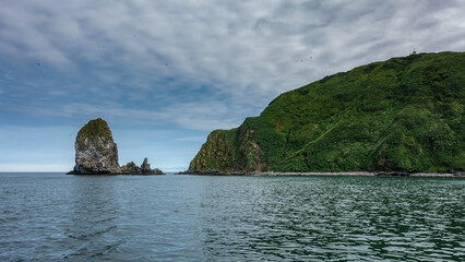 Naklejka premium Islands in the Pacific Ocean against a background of blue sky and clouds. Steep rocky slopes. Green vegetation on the hills. Kamchatka. Avacha Bay