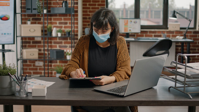 Entrepreneur working with charts papers and laptop to plan business project. Woman wearing face mask, holding clipboard with files to check data analysis for marketing presentation.