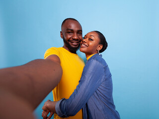 Obraz premium POV of man holding camera and woman hugging him in studio, taking pictures. Romantic couple sharing affection and love together, embracing over blue background. Happy people in relationship.