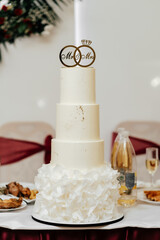 Wedding cake. Elegant, tall wedding cake, four floors with beige mastic and cream with golden breezes with wedding rings on the table