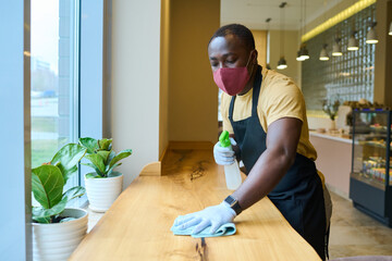 African waiter cleaning the table with spray disinfectant on table in restaurant during pandemic