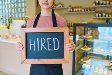 Close-up of young woman in apron holding board with hired sign standing in cafe, she getting new...