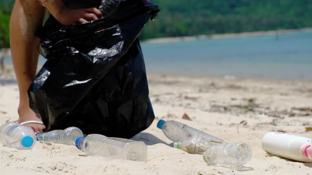 A woman volunteer picking up plastic bottles into a black garbage bag on a sandy beach, environmental protection and ocean cleaning and saving concept.	