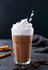 Hot chocolate with whipped cream, tube and cinnamon in a tall glass on a dark background. Front...