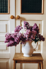 bouquet of purple and white lilacs in a white ceramic vase. lilac flowers in a vase on a wooden shelf. a bouquet of flowers on the background of the doors