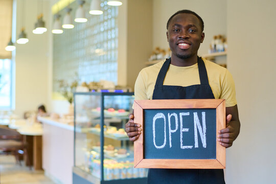 Smiling African waiter in apron holding board with open sign and welcoming guests to new cafe