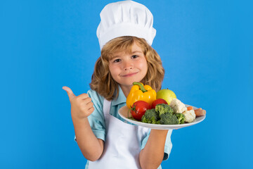 Child chef hold plate with vegetables isolated on blue. Funny little kid chef cook wearing uniform cook cap and apron cooked food in the kitchen.