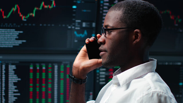 African-American trader talk on smartphone standing over multiple monitors with charts and data