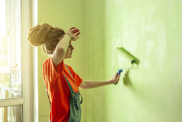 Woman with dreadlocks, in an orange t-shirt and denim jumpsuit is painting the wall with a roller in green color. Concept of repair, renovation of the new apartment