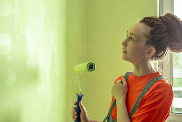 Woman with dreadlocks, in an orange t-shirt and denim jumpsuit is painting the wall with a roller in green color. Concept of repair, renovation of the new apartment. Copy space
