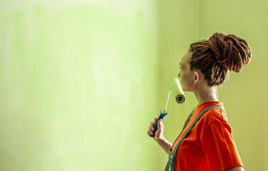 Woman with dreadlocks, in an orange t-shirt and denim jumpsuit is painting the wall with a roller in green color. Concept of repair, renovation of the new apartment. Copy space