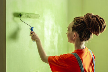 Woman with dreadlocks, in an orange t-shirt and denim jumpsuit is painting the wall with a roller in green color. Concept of repair, renovation of the new apartment