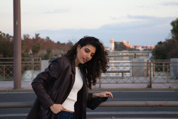 Young and beautiful woman with dark and curly hair is sightseeing in Seville. In the background part of the Mediterranean style European city in the golden hour. Tourism and holidays concept. - obrazy, fototapety, plakaty