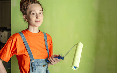 Woman painter with dreadlocks, in an orange T-shirt and a denim jumpsuit make repairs in the apartment and paints the wall green with a roller