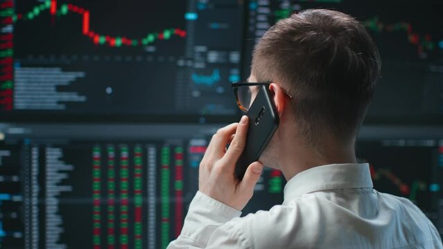 Back view of trader working with multiple computer screens full of charts and data talking on phone