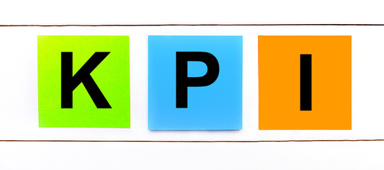 On a white wooden background there are three colorful bright stickers with text KPI Key Performance Indicator