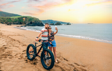 Fototapeta premium Young couple with fat bike taking a selfie with the mobile on the beach