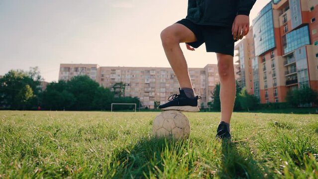 Guy in stadium. Boy stands on grass and keeps his foot on old soccer ball close up. Outdoor sports. Football player in sneakers and shorts. Sports game with ball. Kick ball. Medium plan. Zoom out