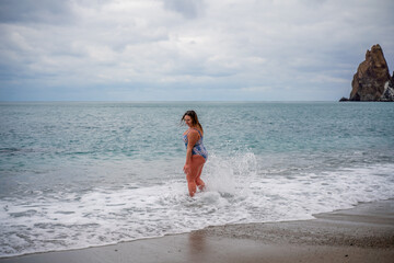 A plump woman in a bathing suit enters the water during the surf. Alone on the beach, Gray sky in the clouds, swimming in winter.