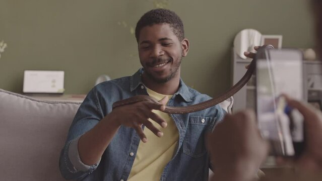 Medium closeup with slowmo of Black woman taking picture on smartphone of her boyfriend holding brown rat snake in hands and smiling, sitting on sofa in their living room