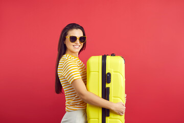 Portrait of smiling young woman in sunglasses hold suitcase isolated on red studio background. Happy female traveler with baggage ready for summer vacation. Tourism and travel concept.