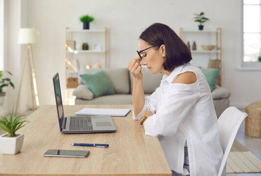 Stressed Exhausted Woman Sitting In Front Of Laptop Computer Screen With Her Dry Irritated Eyes Closed. Side View Of Tired Middle Aged Lady With Eye Strain Sitting At Work Desk And Rubbing Nose Bridge