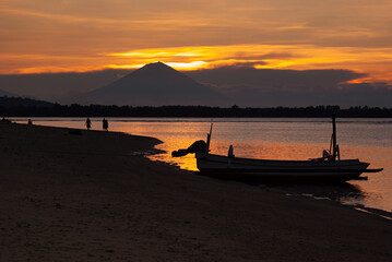 Majestic sunset on sea - golden sky, last sunbeams, dark volcano, black silhouette of fishing boat and walking people on beach, calm orange water. Tropical landscape in Indonesia, travel in asia.