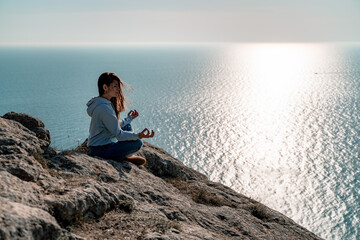Woman tourist enjoying the sunset over the sea mountain landscape. Sits outdoors on a rock above the sea. She is wearing jeans and a blue hoodie. Healthy lifestyle, harmony and meditation