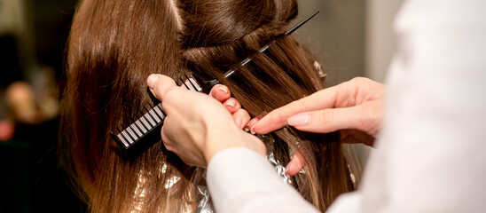 Hairdresser's hands prepare brown hair for dyeing with a comb and foil in a beauty salon, close up
