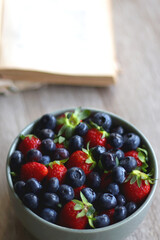 Bowl of strawberries and blueberries, open books with reading glasses, plate of chocolate chip cookies and basket of apples one the table. Selective focus.
