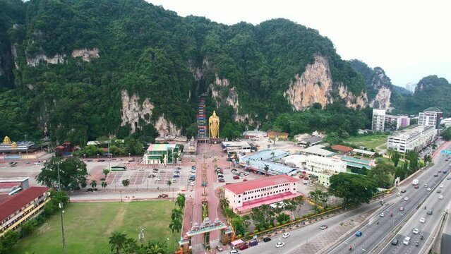 wide aerial circling the entrance to the famous Batu Caves in Kuala Lumpur Malaysia on a cloudy afternoon with highway traffic nearby and large limestone mountains in the distance.