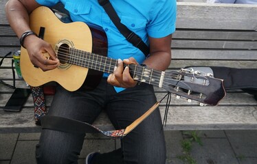Mann in hellblauem Hemd und schwarzer Jeans sitz auf Holzbank und spielt Gitarre in Stadt im...