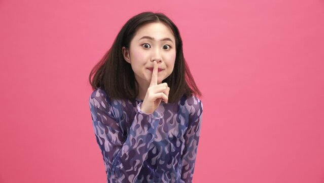Cheerful Asian woman looking around and showing silence gesture at the camera in the pink studio