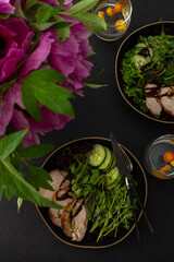 Hearty delicious lunch, fresh salad with cucumbers and microgreens of peas and with stewed pork, top view of the dark table with food and a vase of flowers
