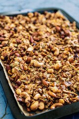 Freshly made homemade granola on a baking tray. Selective focus on nuts in the front, blurred nuts background. Mixed nuts, raisins, coconut flakes, chia and coconut oil. Blue wooden background. 