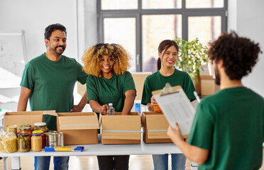 charity, donation and volunteering concept - international group of happy smiling volunteers packing food in boxes according to list on clipboard at distribution or refugee assistance center