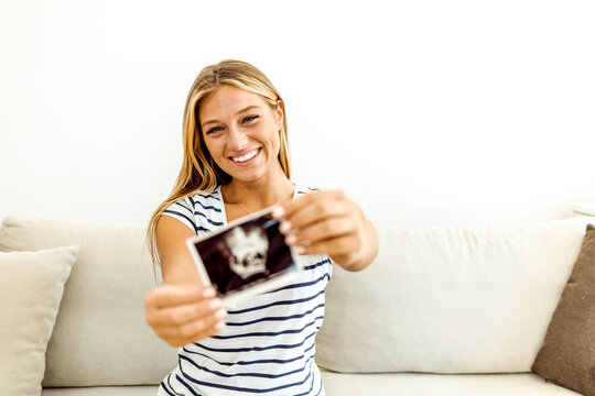 Young smiling pregnant woman holds an ultrasound picture with her baby. Shot of a woman sitting with a sonogram on her bed. Young, pregnant woman examining her ultrasound. Pregnancy and people concept
