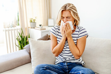 Shot of an attractive young woman suffering with the flu while sitting on her bed at home. The sick woman, holding a handkerchief, sneezing and feeling freezing, sitting on bed at home. Health problem - obrazy, fototapety, plakaty