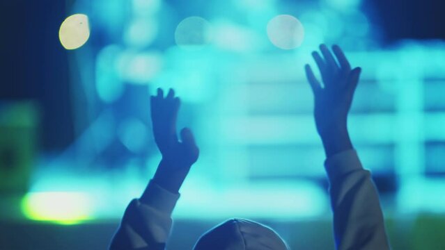 Young man claps palm with hands raised above head having fun at an open air concert, back view. Man happily claps his hands to beat of music at an outdoor party