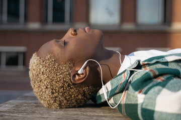 Close up portrait of stylish African American man relaxing, listening music with closed eyes lying on the street, selective focus